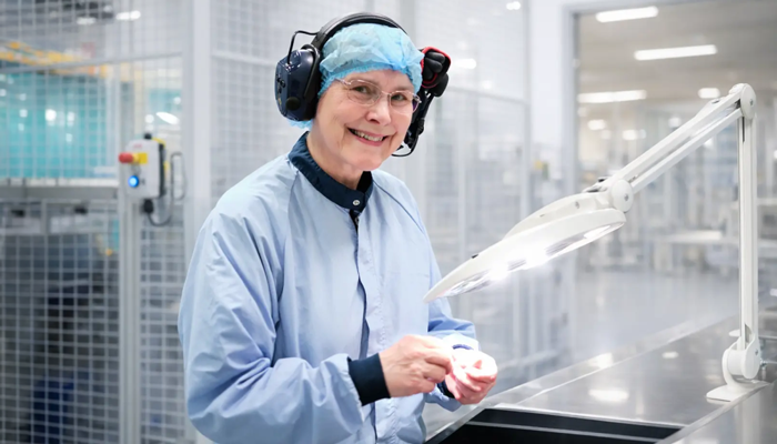 A smiling employee in cleanroom clothing, hair cover, safety glasses, and hearing protection works at a brightly lit station in a clean manufacturing environment, holding a small component under a task lamp.