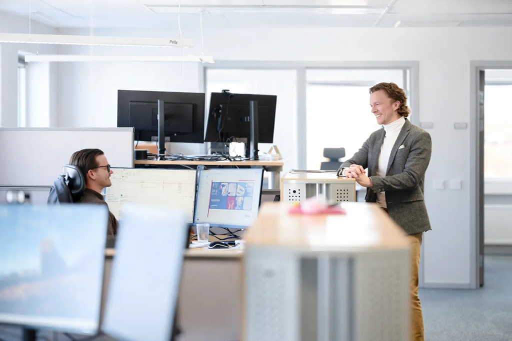 Two colleagues talk and smile in a modern open-plan office with several computer monitors and desks in the foreground. One person is seated at a workstation while the other stands beside a cabinet in a bright, professional workspace.