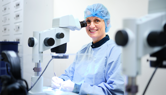 A smiling employee in cleanroom clothing, hair cover, glasses, and gloves sits beside a microscope in a bright laboratory environment, holding a small tool while performing detailed inspection work.