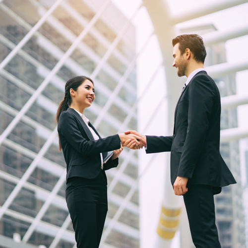 A woman and a man shaking hands in front of office buildings.