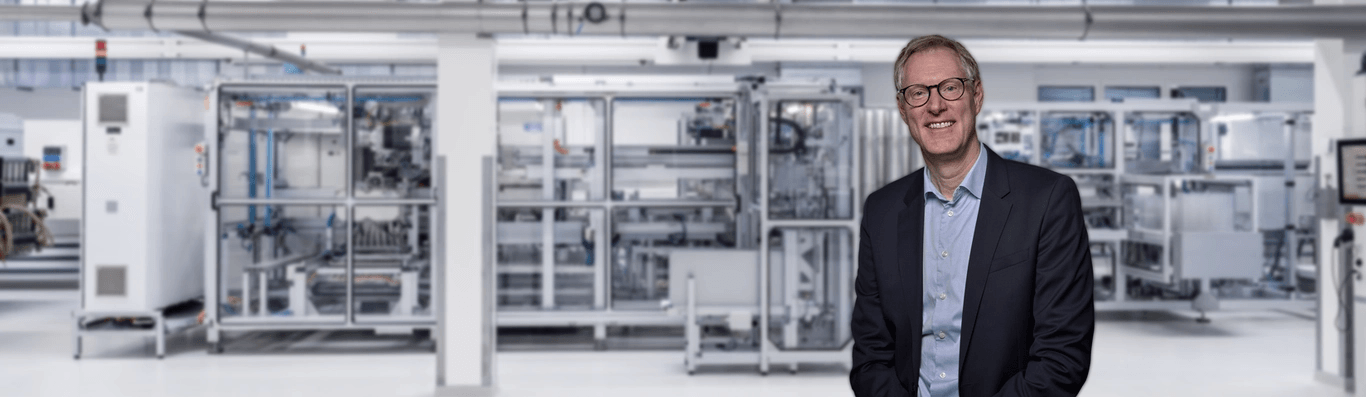 Smiling man in suit and glasses in a modern, brightly lit lab or factory with intricate white and silver machinery.