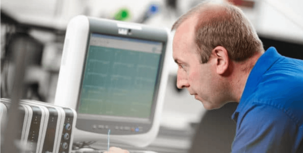 Man in blue shirt with receding hairline intently views graphs on a computer monitor next to stacked equipment.