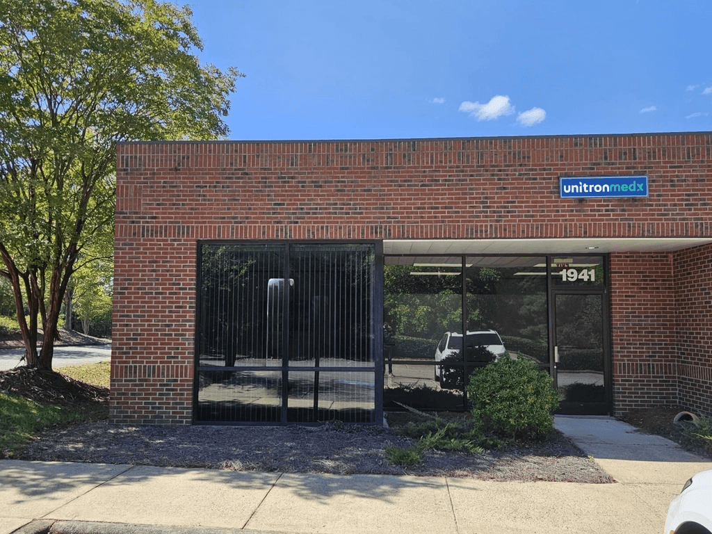 Unitron in US. Single-story brick building with two large dark windows. Trees and clear blue sky.