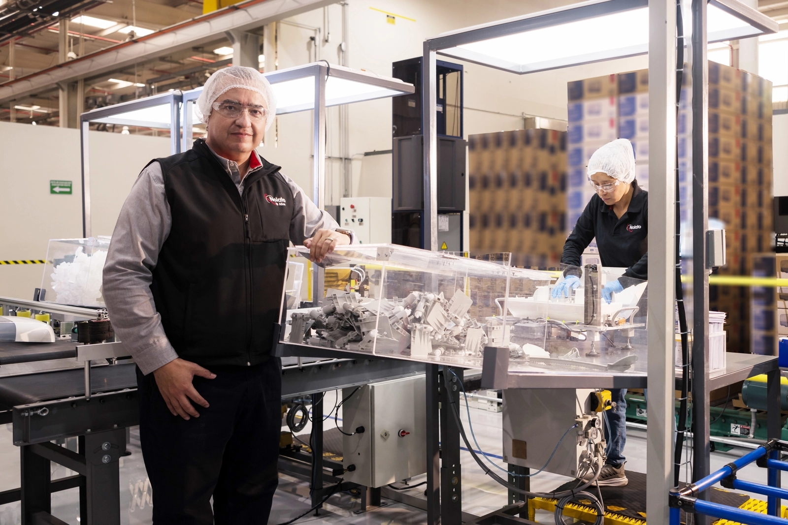 Man in hairnet and vest leaning on packaging machine in a clean factory, with another worker and stacked boxes in the background.