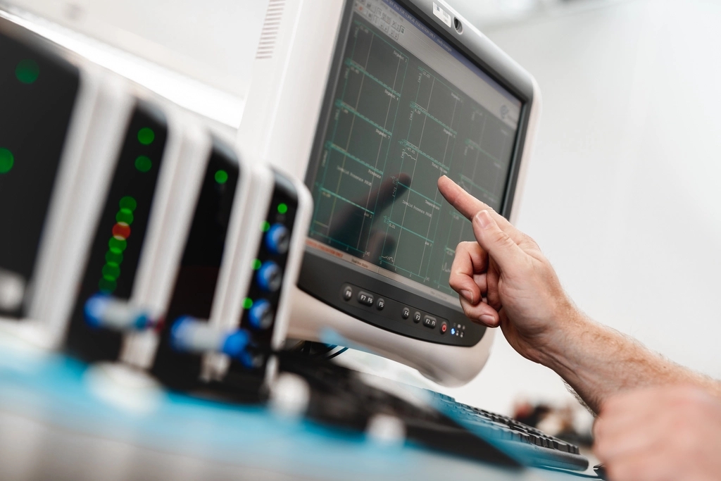 Hand pointing at a monitor displaying a waveform graph, next to a row of electronic lab equipment with lights.