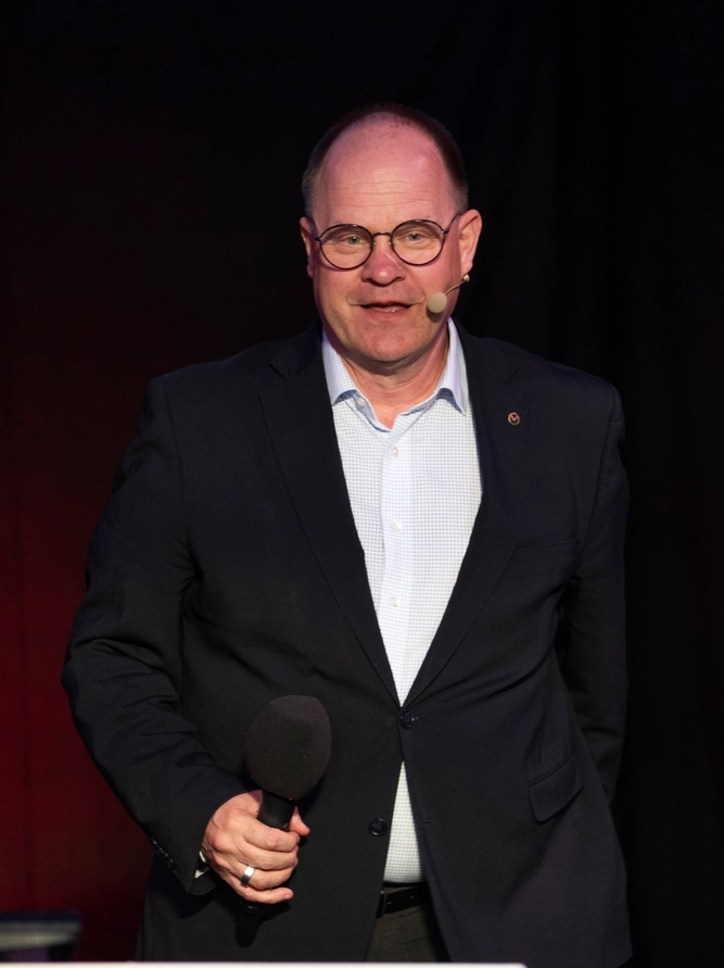 Man in a dark suit, glasses, and earpiece, holding a microphone on a stage.