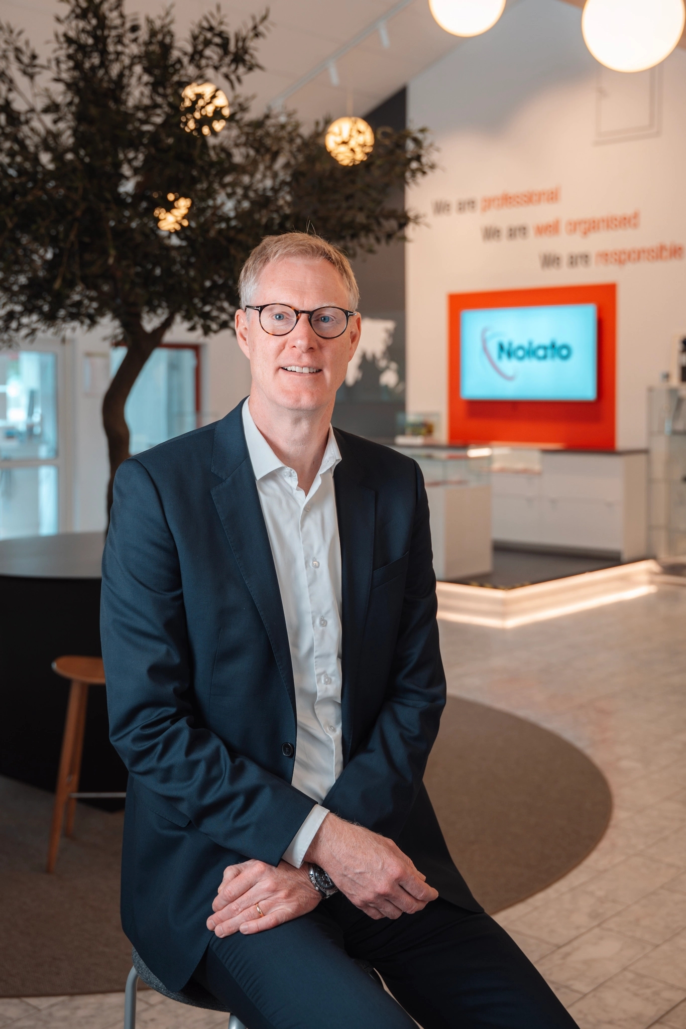 Man in suit and glasses sitting in a modern office lobby with a "Nolato" screen and "We are professional" text on the wall.