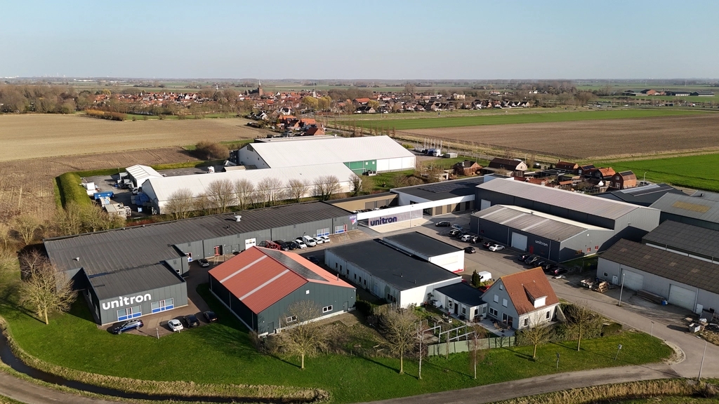 Multiple industrial and commercial buildings of various sizes and colors, nestled between green fields and a distant village.