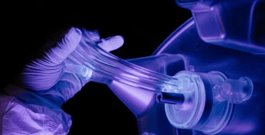 Gloved hand connects glowing clear tubes to a transparent medical device, under purple light.