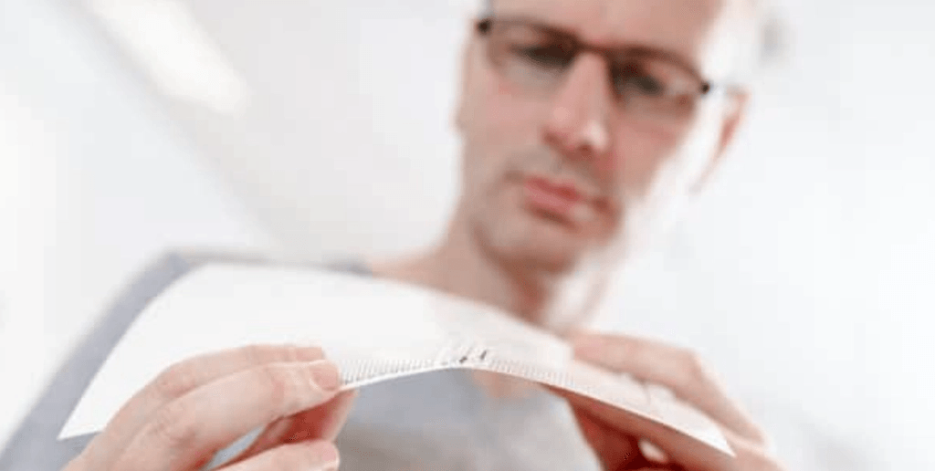 Hands in foreground bend a thin, flexible white sheet; man with glasses observes in the blurred background.