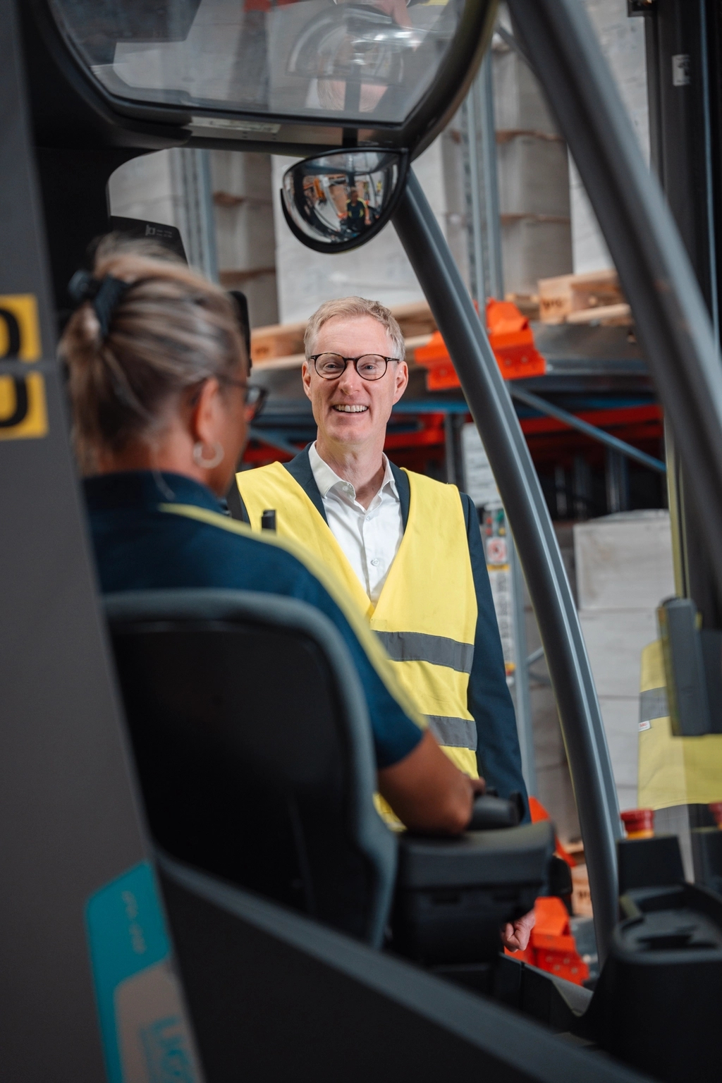 Smiling man in high-vis vest talks to a forklift operator in a warehouse.