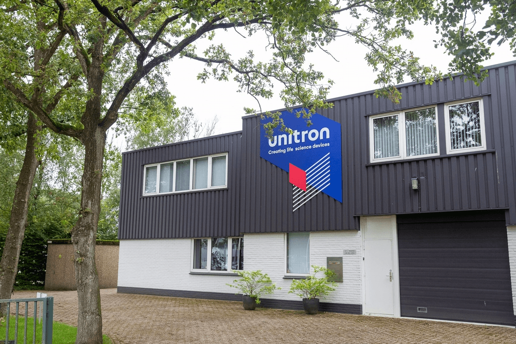Unitron building in Hengelo with dark gray and white facade, blue sign with 'unitron' and geometric logo. Trees line the drive.