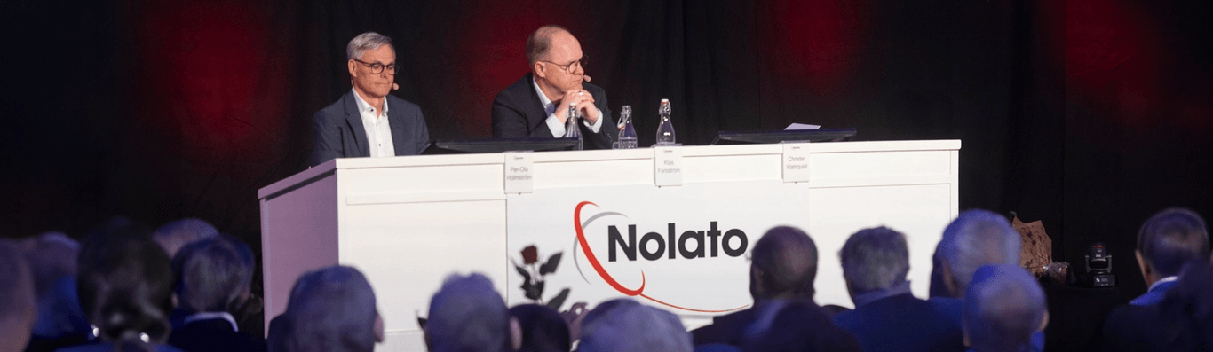 Two men seated behind a white table with "Nolato" logo, facing an audience. Dark background with red stage lights.
