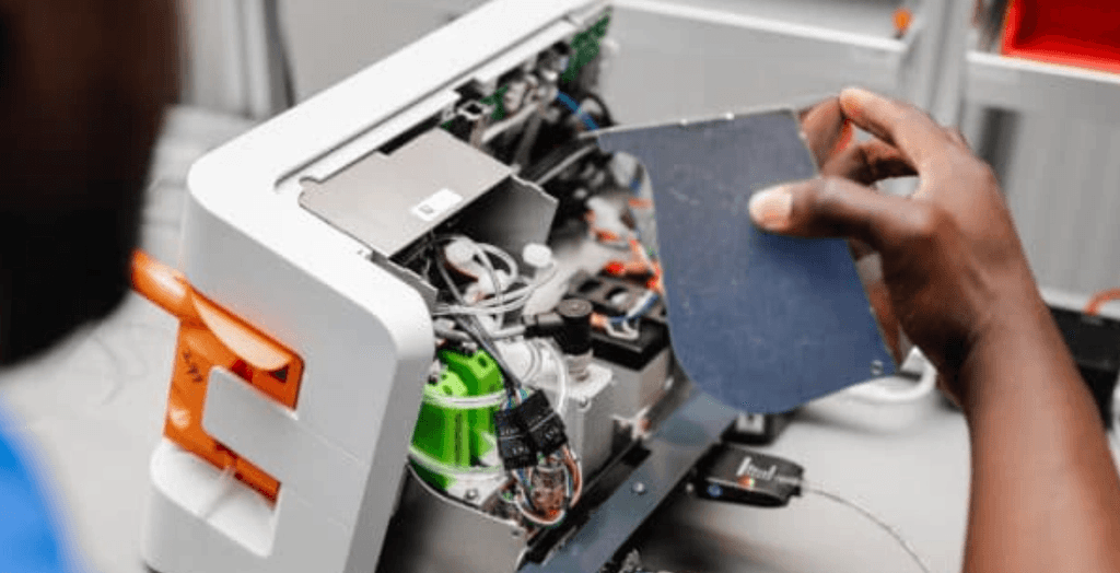 Hand holds a component over an open white machine, revealing complex internal electronics, wires, and circuit boards.