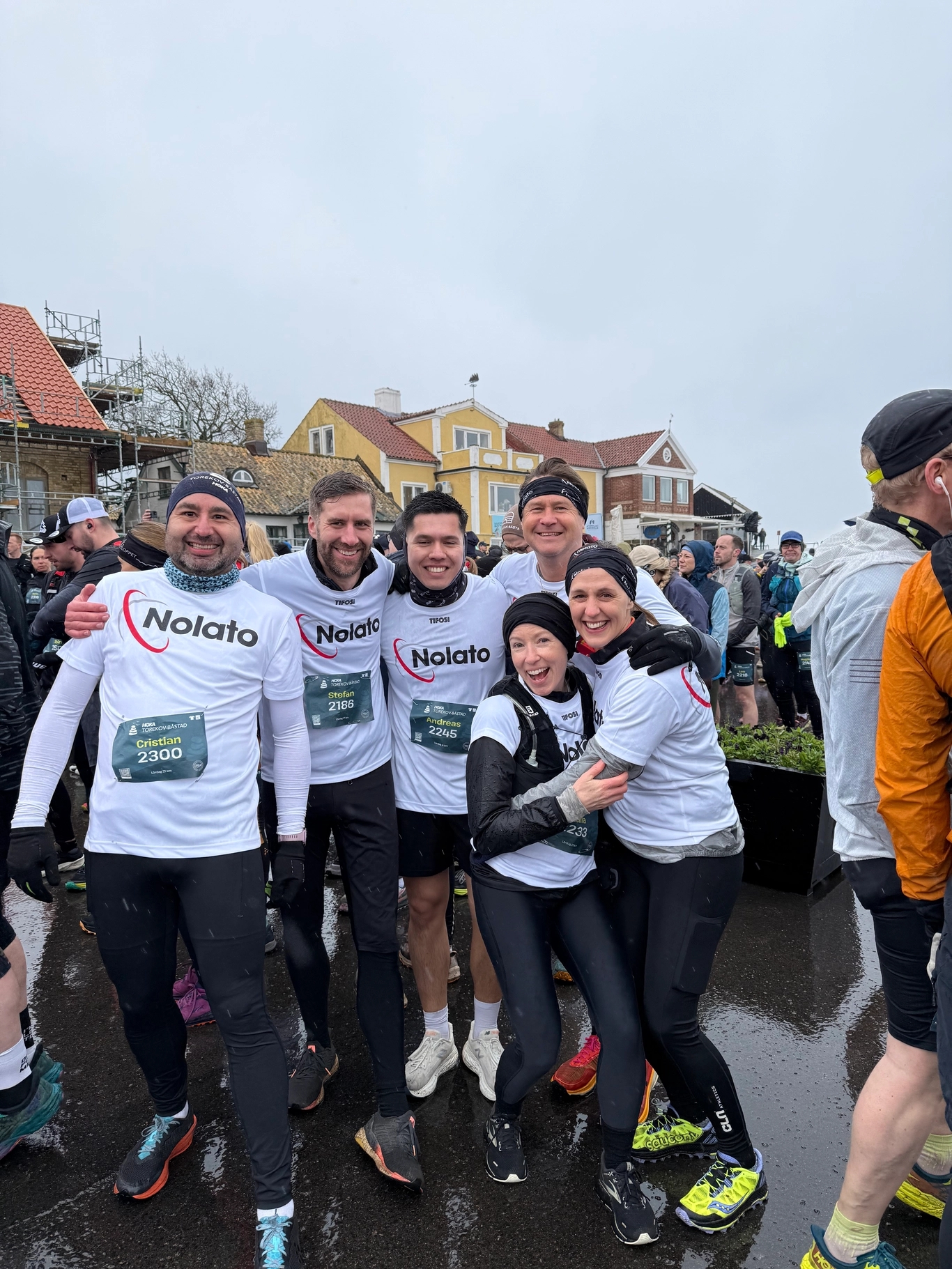 Group of six smiling people in white "Nolato" shirts and black athletic wear posing at an outdoor race event.